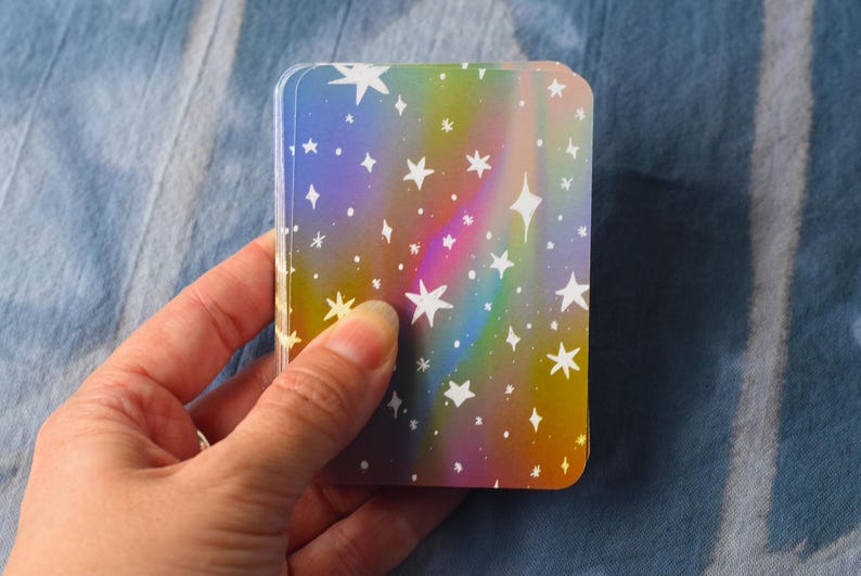 Pode incluir: Um baralho de cartas retangulares com um fundo hologr&aacute;fico de cores do arco-&iacute;ris e um padr&atilde;o repetitivo de estrelas brancas desenhadas &agrave; m&atilde;o de v&aacute;rios tamanhos espalhadas pela superf&iacute;cie de cada carta no monte.