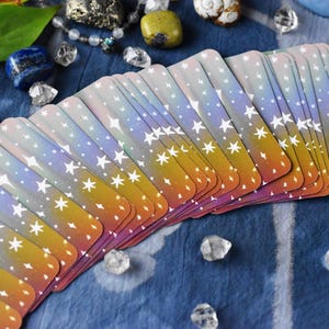 Pode incluir: Um baralho de cartas de tarot disposto em leque, com um design em gradiente que transita de um violeta suave no topo para um laranja quente na base, decorado com um padr&atilde;o de estrelas brancas de quatro pontas de v&aacute;rios tamanhos.