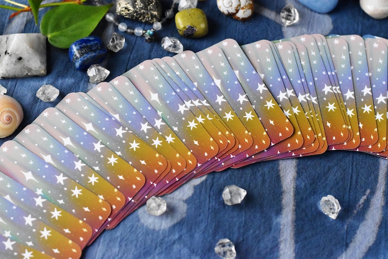 Pode incluir: Um baralho de cartas de tarot disposto em leque, com um design em gradiente que transita de um violeta suave no topo para um laranja quente na base, decorado com um padr&atilde;o de estrelas brancas de quatro pontas de v&aacute;rios tamanhos.