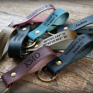 Op de afbeelding: Een set van zes leren sleutelhangers met verschillende gravures. De sleutelhangers zijn in verschillende kleuren, waaronder bruin, zwart, groen en blauw. De gravuren omvatten namen, coördinaten en een zin in een vreemde taal.