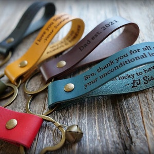 May include: A set of six leather keychains with different colors and engraved messages. The keychains feature a brass key ring and a small bell charm. The messages include "I love you", "Your Sister", "Dad est. 2021", and "Bro, thank you for all of your unconditional love. -Lil Sis".