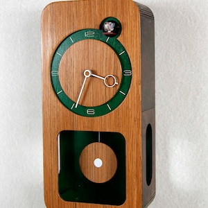 May include: A wooden clock with a green face and a pendulum. The clock has a small window on the top that shows a bird.