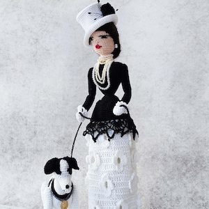 May include: A crocheted doll wearing a black dress and a white top hat, holding a leash attached to a crocheted black and white dog. The doll has a pearl necklace and red lips.