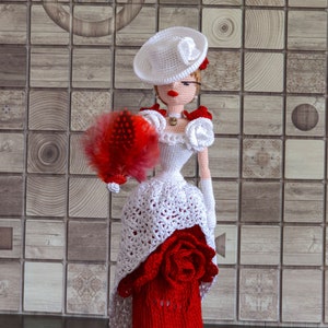 May include: A crocheted doll wearing a white dress with a red rose accent, a white hat, and holding a red feather fan. The doll is standing in front of a tiled background.