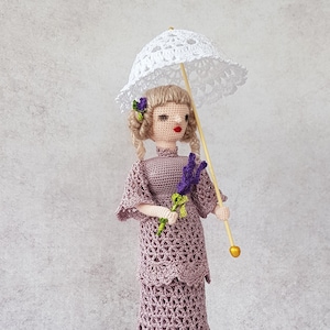 May include: A crocheted doll wearing a lavender dress and a white crocheted hat, holding a bouquet of purple flowers and a white parasol.