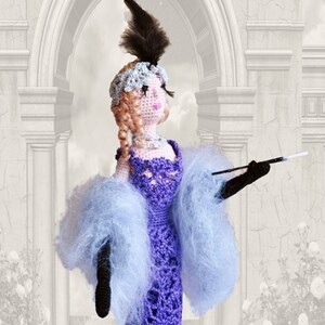 May include: A crocheted doll wearing a purple dress with a blue feather boa and a black feather headpiece. The doll is holding a cigarette in its right hand.