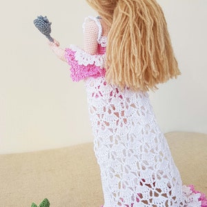 May include: A crocheted doll wearing a white dress with pink trim and a pink bodice. The doll has long blonde hair and is holding a gray crocheted rose.