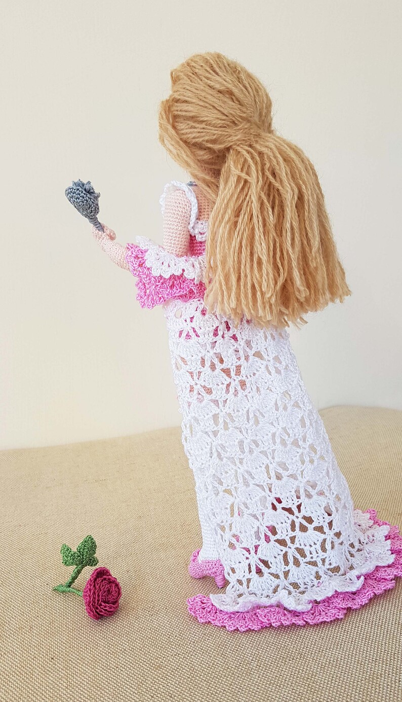 May include: A crocheted doll wearing a white dress with pink trim and a pink bodice. The doll has long blonde hair and is holding a gray crocheted rose.