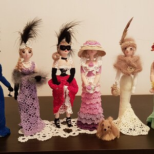 May include: Six crocheted dolls dressed in vintage-inspired outfits, each with a unique hat and accessories. The dolls are standing on a wooden surface with a small crocheted dog in front of them.