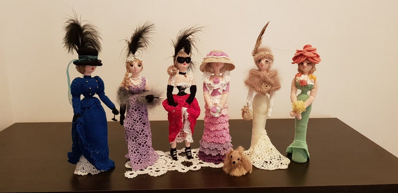 May include: Six crocheted dolls dressed in vintage-inspired outfits, each with a unique hat and accessories. The dolls are standing on a wooden surface with a small crocheted dog in front of them.