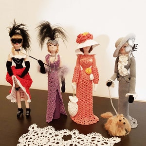 May include: Four crocheted dolls dressed in different outfits. The dolls are standing on a table with a white doily in front of them. The dolls are wearing hats and have different hairstyles. One doll is holding a cane, another is holding a purse, and another is holding a leash with a small crocheted dog.
