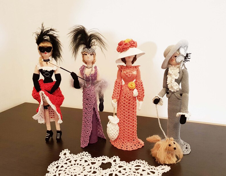 May include: Four crocheted dolls dressed in different outfits. The dolls are standing on a table with a white doily in front of them. The dolls are wearing hats and have different hairstyles. One doll is holding a cane, another is holding a purse, and another is holding a leash with a small crocheted dog.