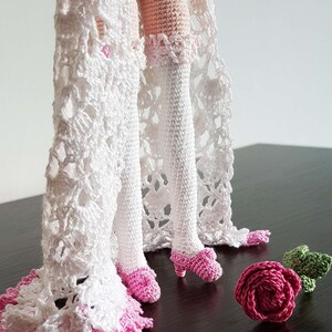May include: Crocheted doll with white lace dress and pink shoes. The doll is standing on a dark surface with a pink crocheted rose and a green crocheted leaf.