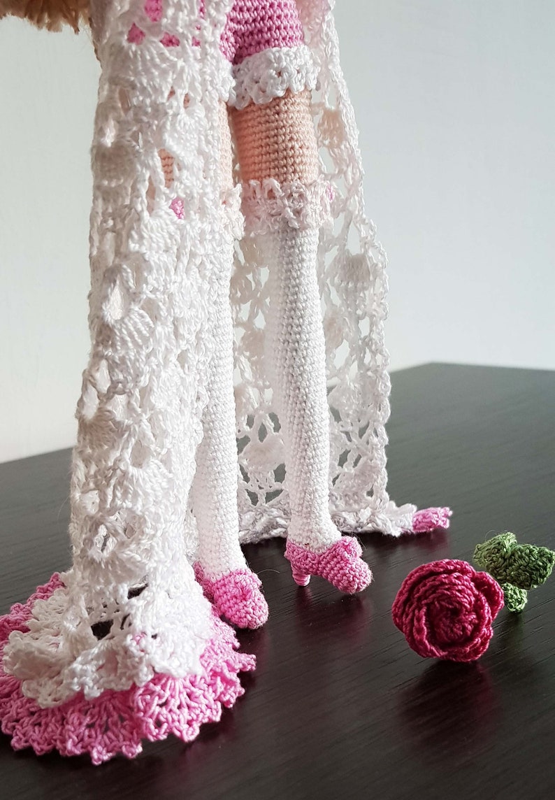 May include: Crocheted doll with white lace dress and pink shoes. The doll is standing on a dark surface with a pink crocheted rose and a green crocheted leaf.