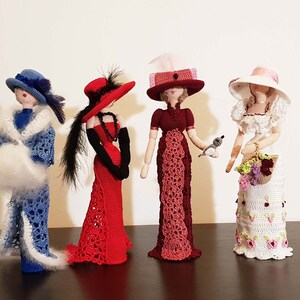 May include: Four crocheted dolls dressed in vintage clothing. The dolls are wearing hats and holding accessories. The dolls are standing on a wooden surface.