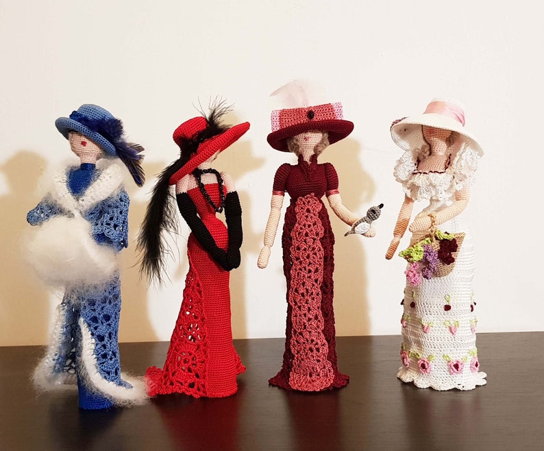 May include: Four crocheted dolls dressed in vintage clothing. The dolls are wearing hats and holding accessories. The dolls are standing on a wooden surface.
