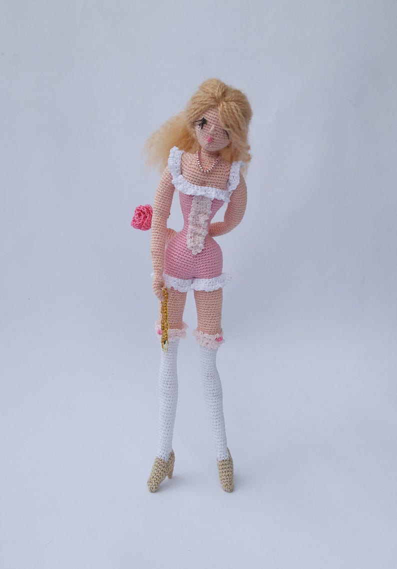 May include: A crocheted doll with blonde hair, wearing a pink and white dress, white stockings, and brown shoes. The doll is holding a pink rose.