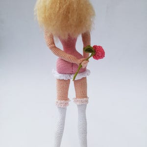 May include: A crocheted doll with blonde hair, wearing a pink dress and white stockings, holding a pink flower.