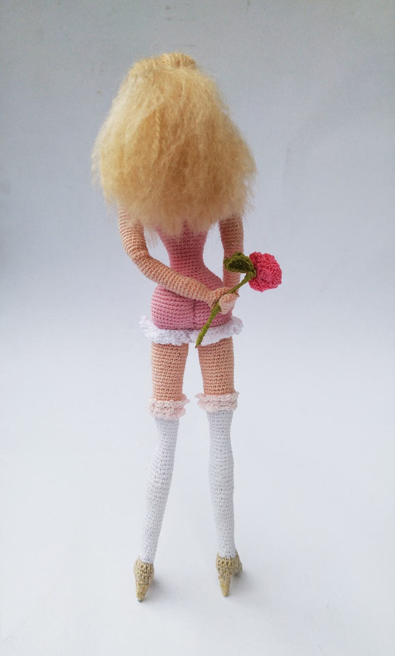 May include: A crocheted doll with blonde hair, wearing a pink dress and white stockings, holding a pink flower.