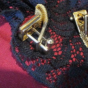 May include: Close-up of two gold-toned cufflinks with rectangular red stone insets. The cufflinks are set against a dark background with a red and black lace pattern. The cufflinks have a classic design.