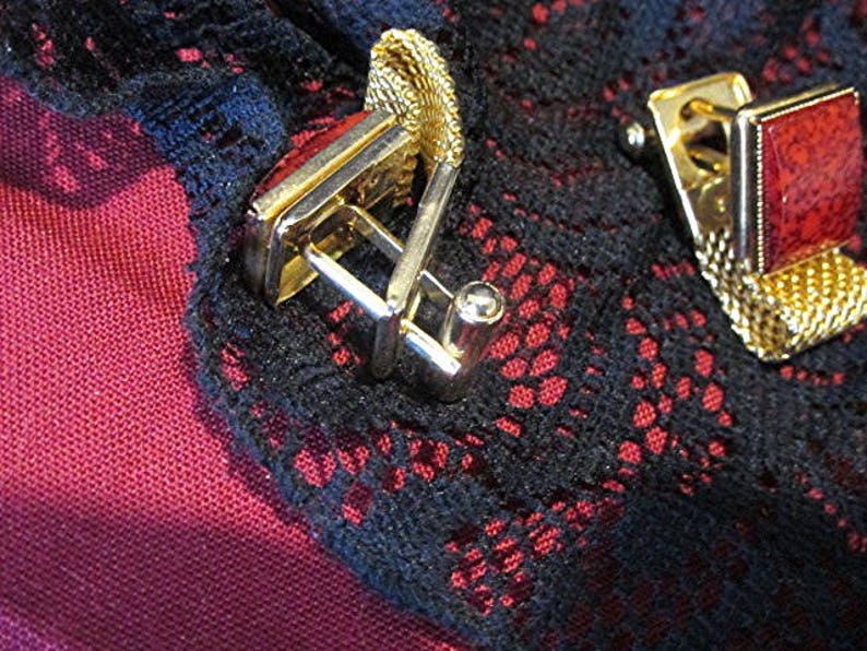 May include: Close-up of two gold-toned cufflinks with rectangular red stone insets. The cufflinks are set against a dark background with a red and black lace pattern. The cufflinks have a classic design.