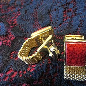 May include: A pair of gold-toned cufflinks with a rectangular red stone inlay. One cufflink features a mesh band, while the other has a chain fringe. The cufflinks are set against a dark, patterned background.