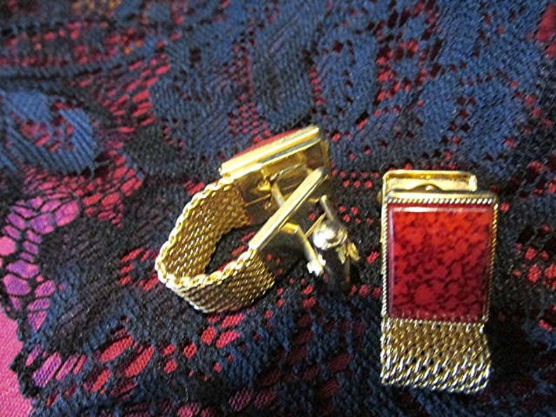 May include: A pair of gold-toned cufflinks with a rectangular red stone inlay. One cufflink features a mesh band, while the other has a chain fringe. The cufflinks are set against a dark, patterned background.