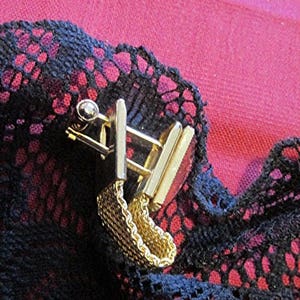 May include: Close-up of a gold-toned metal cufflink set with a chain detail. The cufflinks are rectangular with a small, round accent. The background is a dark blue lace fabric over a red surface.