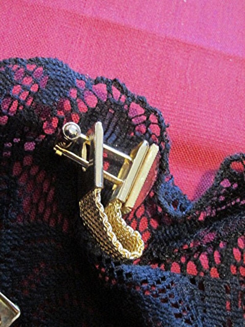 May include: Close-up of a gold-toned metal cufflink set with a chain detail. The cufflinks are rectangular with a small, round accent. The background is a dark blue lace fabric over a red surface.