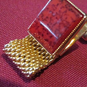 May include: A gold-toned cufflink with a square, reddish-brown stone set in a gold frame. The cufflink features a mesh-like gold chain detail. The background is a textured, maroon fabric.