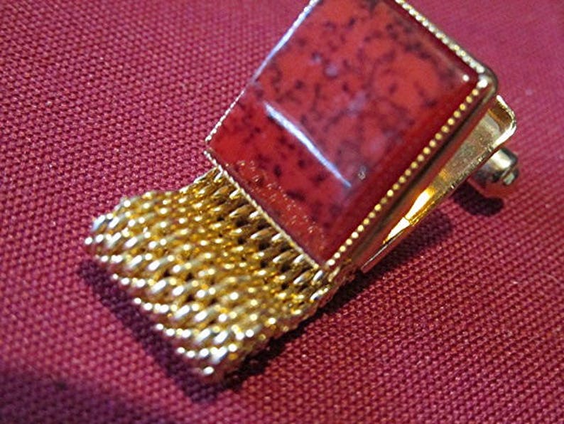 May include: A gold-toned cufflink with a square, reddish-brown stone set in a gold frame. The cufflink features a mesh-like gold chain detail. The background is a textured, maroon fabric.
