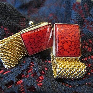 May include: A pair of gold-toned cufflinks with rectangular red speckled insets. The cufflinks have a mesh design and are set against a dark, textured background.