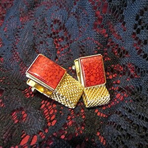 May include: A pair of vintage cufflinks with rectangular red stone insets and gold-toned mesh detailing. The cufflinks are set against a dark background with a lace-like pattern, creating a striking contrast.