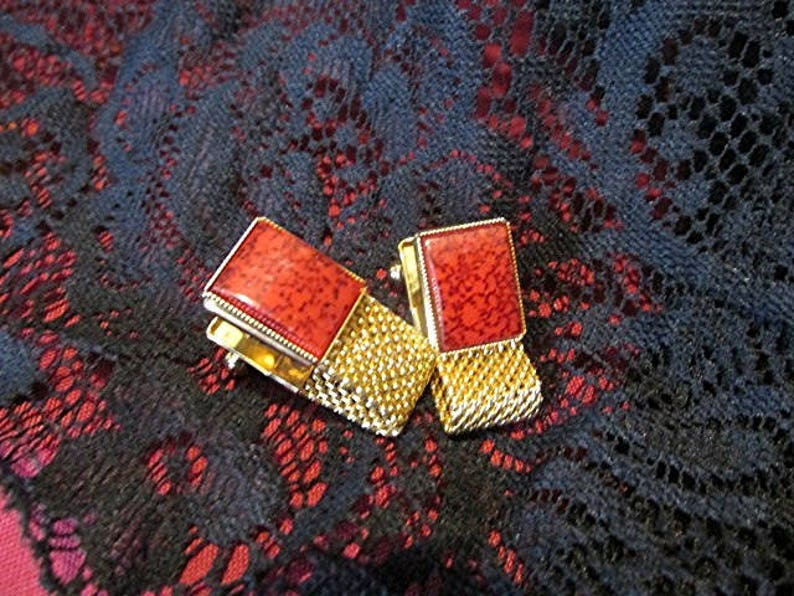 May include: A pair of vintage cufflinks with rectangular red stone insets and gold-toned mesh detailing. The cufflinks are set against a dark background with a lace-like pattern, creating a striking contrast.