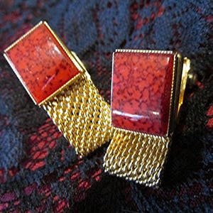 May include: A pair of gold-toned cufflinks with rectangular red stone faces. The red stones have a speckled pattern. The cufflinks have a gold chain detail. The background is a dark, patterned fabric.