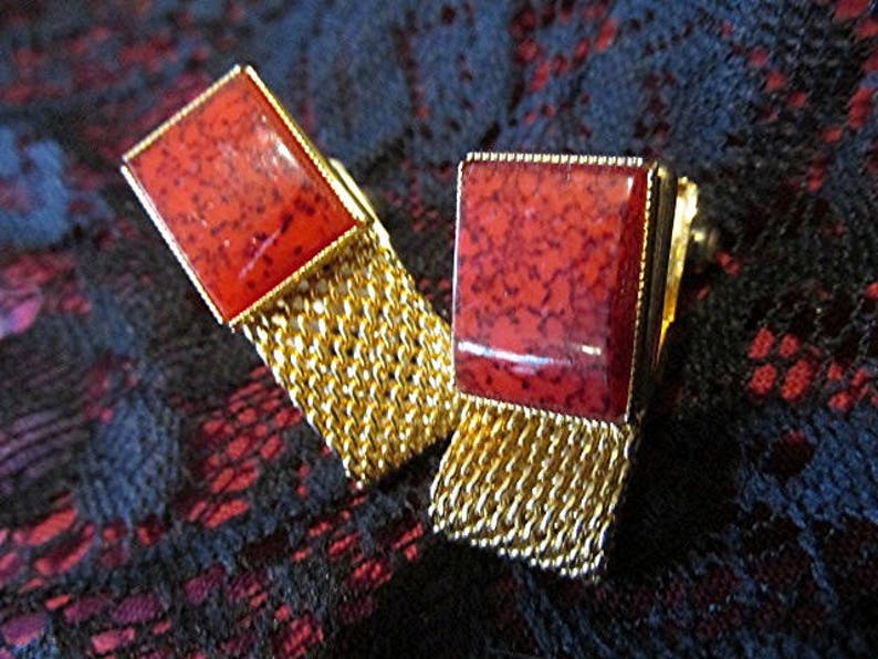 May include: A pair of gold-toned cufflinks with rectangular red stone faces. The red stones have a speckled pattern. The cufflinks have a gold chain detail. The background is a dark, patterned fabric.