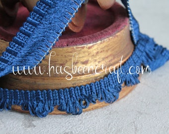 May include: Blue fringe trim with a woven texture. The fringe is approximately 1 inch wide and has a subtle sheen. The trim is wrapped around a wooden spool.