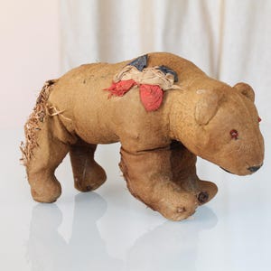 Peut inclure: Un ours en peluche brun vintage avec des yeux rouges et des morceaux de tissu sur le dos. L'ours a une apparence usée, suggérant qu'il s'agit d'une antiquité ou d'un objet très apprécié. Le jouet est à quatre pattes.