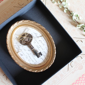 Könnte beinhalten: Ein antiker Schlüssel wird in einem goldenen ovalen Rahmen präsentiert, der auf einem weißen Papier mit Schrift ruht. Der Rahmen befindet sich in einer schwarzen Box, mit einem floralen Zweig und gemustertem Papier im Hintergrund. Der Schlüssel ist mit Bindfaden gebunden.