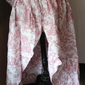May include: A white and red toile skirt with a classic pastoral scene print. The skirt is gathered at the waist and drapes down, showcasing the detailed illustrations. The fabric is lightweight, ideal for historical reenactment or costume.