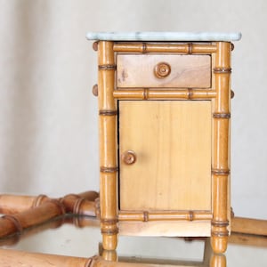 Può includere: Un piccolo comodino antico in stile bambù con piano in marmo. Presenta un singolo cassetto e un'anta, entrambi con pomelli rotondi in legno. La struttura è di colore marrone chiaro.