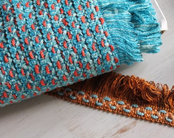 May include: Close-up of a turquoise and orange woven fabric with a fringe. The fabric is folded in half and a brown fringe trim is visible in the foreground.