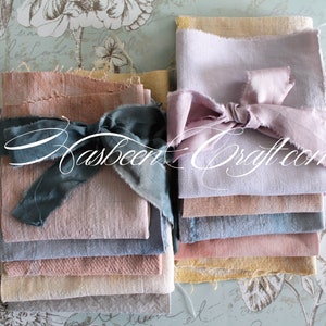Op de afbeelding: Een stapel van acht handgekleurde linnen vierkanten in verschillende tinten roze, blauw en geel. De stof is vastgebonden met een blauw lint en de tekst "Rasbeer Craft.com" is zichtbaar in het midden van de afbeelding.