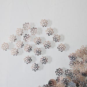 Pode incluir: Uma coleção de pequenos componentes de filigrana metálica em forma de flor. As peças estão dispostas num grupo solto à esquerda e num monte denso à direita, cada uma apresentando um design delicado e aberto com várias pétalas arredondadas.