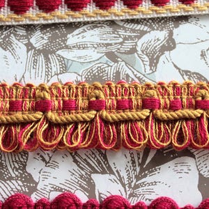 May include: Three distinct decorative fabric trims. The top trim features a red scalloped edge with gold accents. The middle trim is a wide woven band with gold rope detailing and a fringe of red and gold loops. The bottom trim has a red scalloped edge.