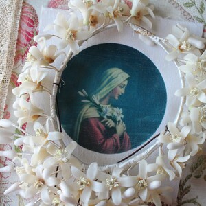 Pode incluir: Imagem religiosa emoldurada de uma mulher com véu branco e túnica vermelha, segurando flores, rodeada por uma grinalda floral branca. A grinalda é adornada com pequenas flores e botões brancos. O fundo inclui renda e tecido floral.