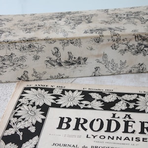 Può includere: Una scatola rettangolare con coperchio incernierato, decorata con un motivo toile de Jouy in bianco e nero. Sotto, una rivista vintage intitolata "LA BRODERIE LYONNAISE". La rivista presenta un bordo floreale in bianco e nero e testo.