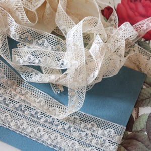Peut inclure: Ruban de dentelle crème, finement tissé avec des motifs délicats, posé sur une surface bleu sarcelle. Le ruban est disposé en spirales et en lignes droites, mettant en valeur son design détaillé. Un imprimé floral vintage avec une rose rouge est en arrière-plan.
