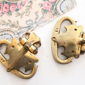 Pode incluir: Duas fivelas antigas de latão com um design curvo, apresentando uma forma de pássaro. As fivelas estão posicionadas em uma superfície branca, com um papel floral no fundo.