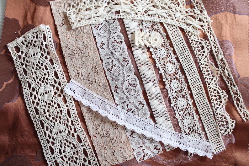 May include: Assortment of white lace trims with various patterns and textures. The lace trims are arranged in rows on a brown fabric background.